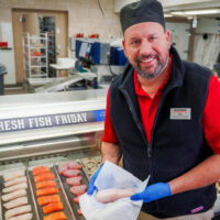 A image of a local harmons butcher showcasing their local meats with the fresh meat and produce section in the background filled with fresh fruits and vegetables at harmons grocery in utah Shop local and eat healthy my local utah